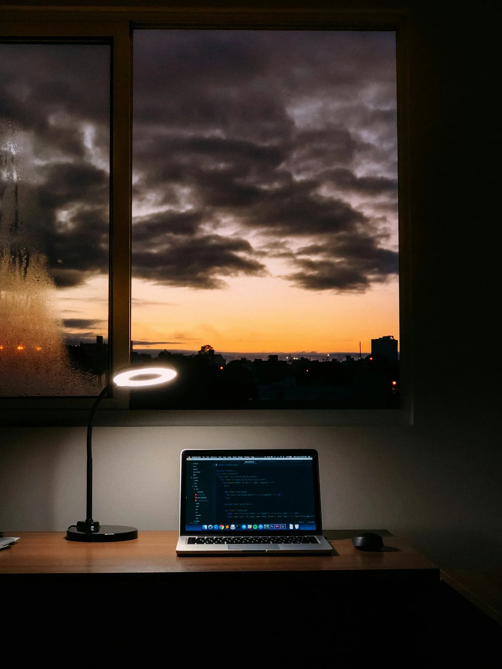 A laptop with code on screen on a desk by a window at dusk